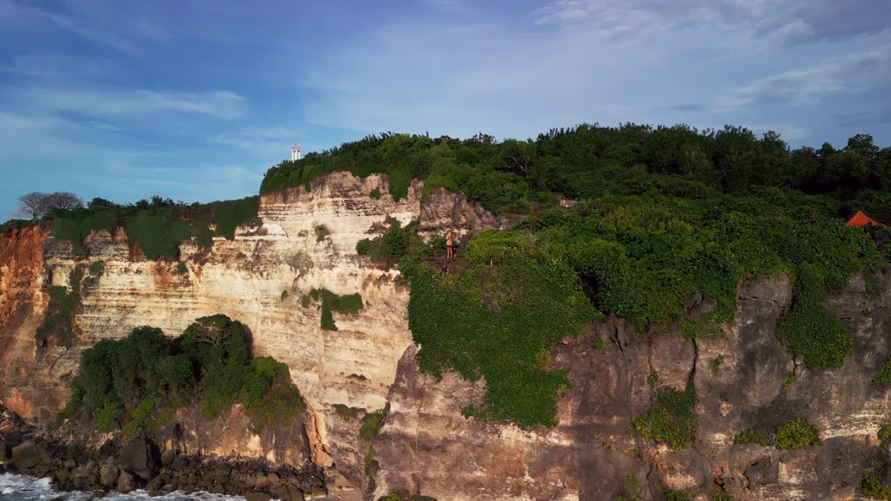 Aerial shot of a person positioned on a rocky cliff along Balis coastline, surrounded by crashing turquoise waves and lush greenery, highlighting the raw scenic power of the island’s coastal landscape