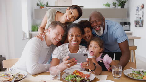 multi-generation mixed race family posing for selfie as they eat meal around table at home together