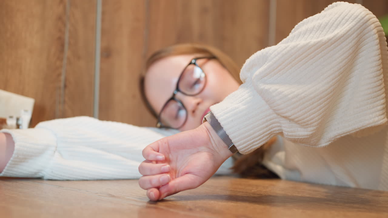 student in white sweater rests head on wooden table while arm stretches forward to check time, capturing quiet reflective moment with minimal background, napkin holder, and subtle indoor lighting