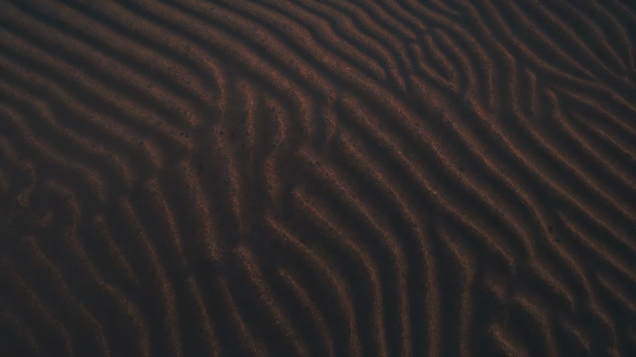 Shifting camera documenting textured sand ripples at beach, low sun highlighting depressed patch
