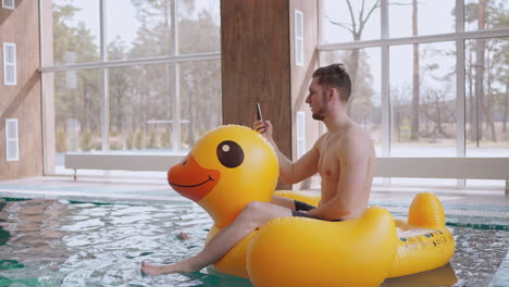 Un-Hombre-Alegre-Llama-Por-Video-En-Un-Teléfono-Móvil-Desde-Una-Piscina-Moderna-Montando-Un-Divertido-Círculo-Inflable