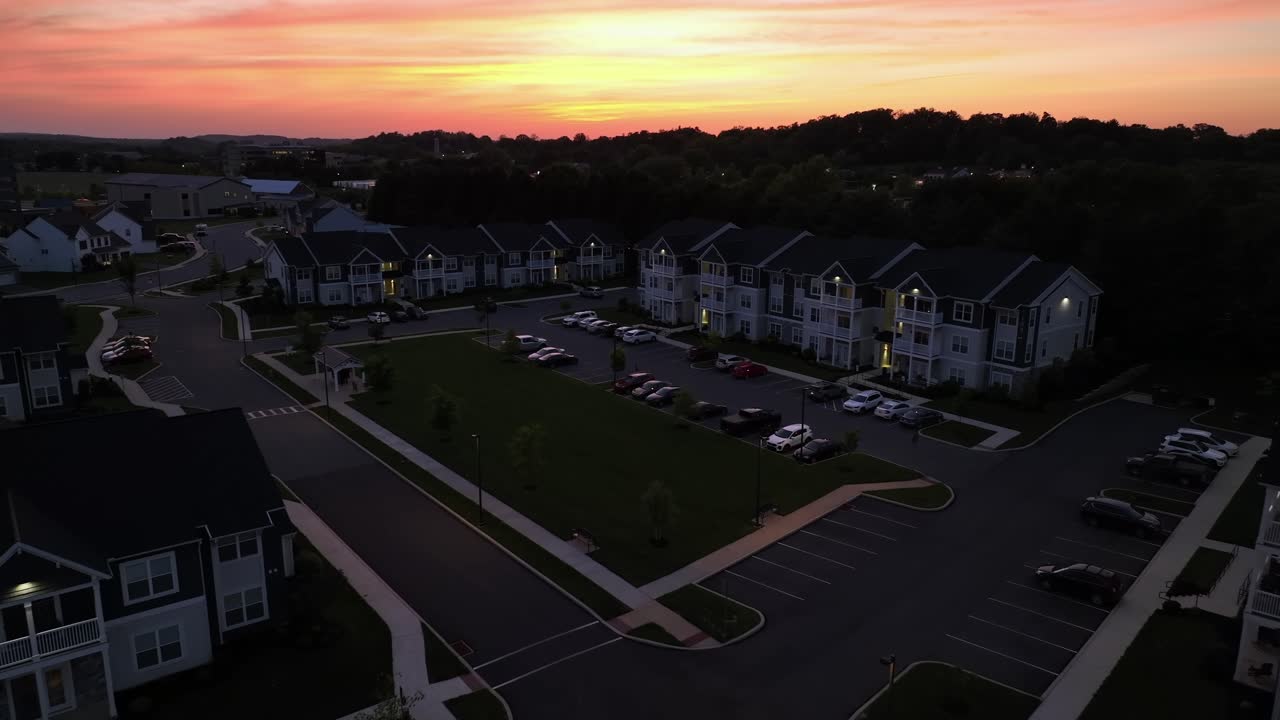 Elegant townhouses with modern designs line the street at sunset. Parking cars in center of neighborhood. Aerial approaching shot. Dusk scene in suburbia of America. Wide shot.