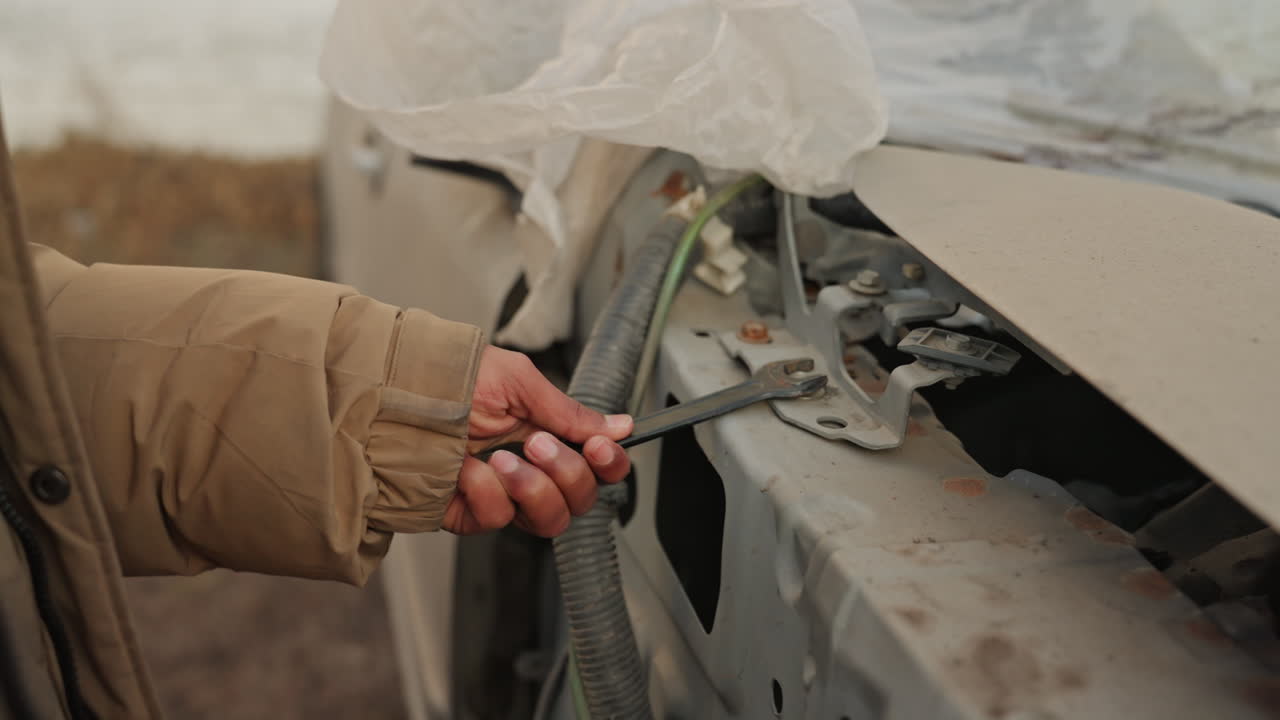 Closeup hand repairing car panel, brown jacket sleeve, rusted metal edges, pliers manipulating clip and cable, stepbystep repair action, overcast outdoor setting with gritty salvage mood