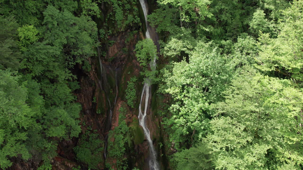 Drone footage of tall waterfall cascading through rocks and lush green forest in summer daylight