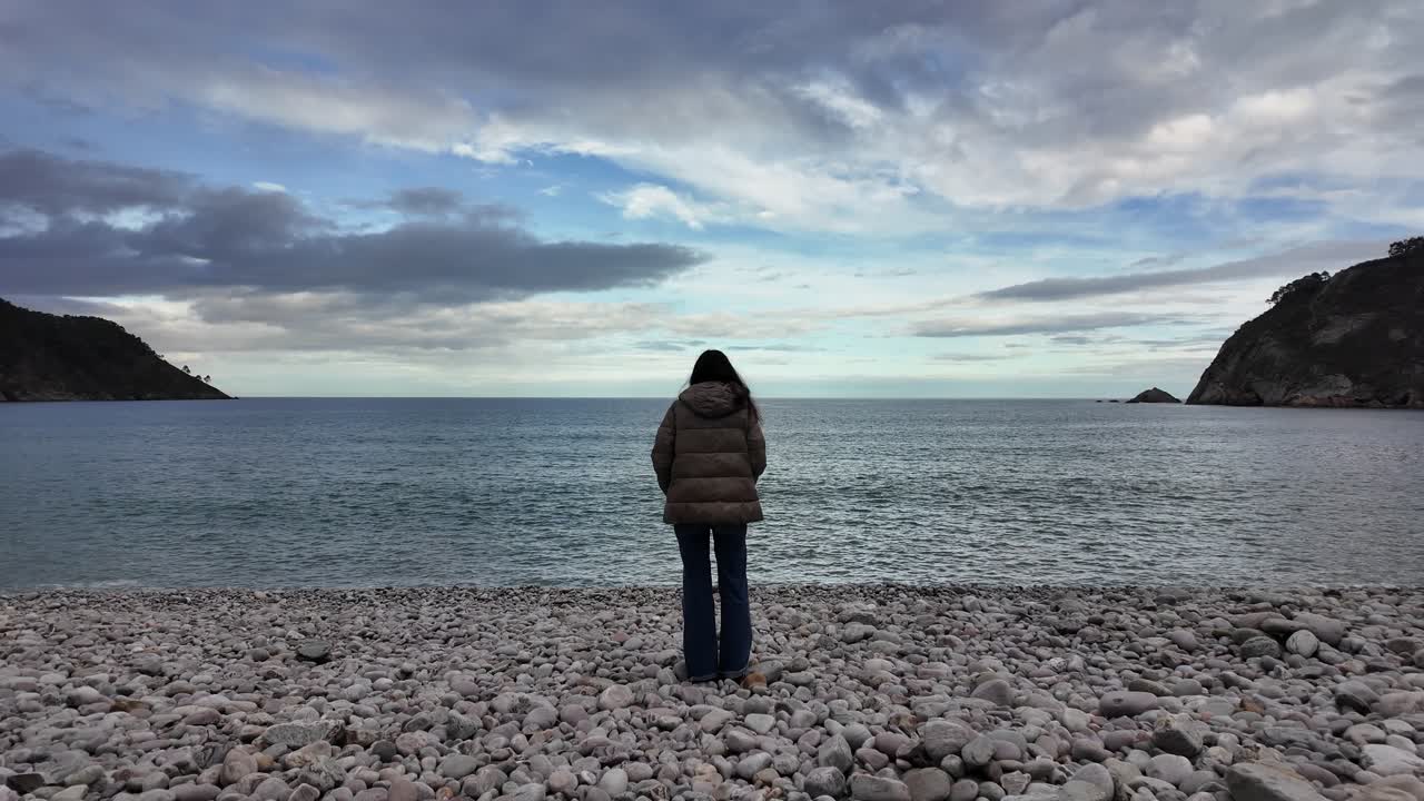 A person stands alone on a pebble beach, facing the calm sea between two headlands