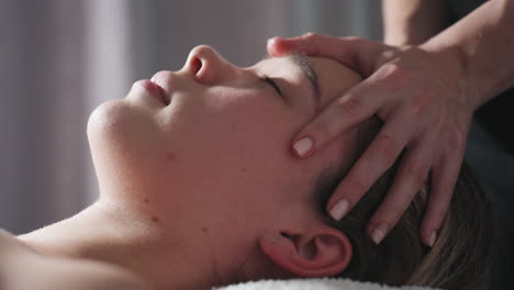 close up of woman lying down with eyes closed while therapist gently massages forehead and scalp, creating soothing wellness ambiance with soft lighting promoting tranquility and comfort