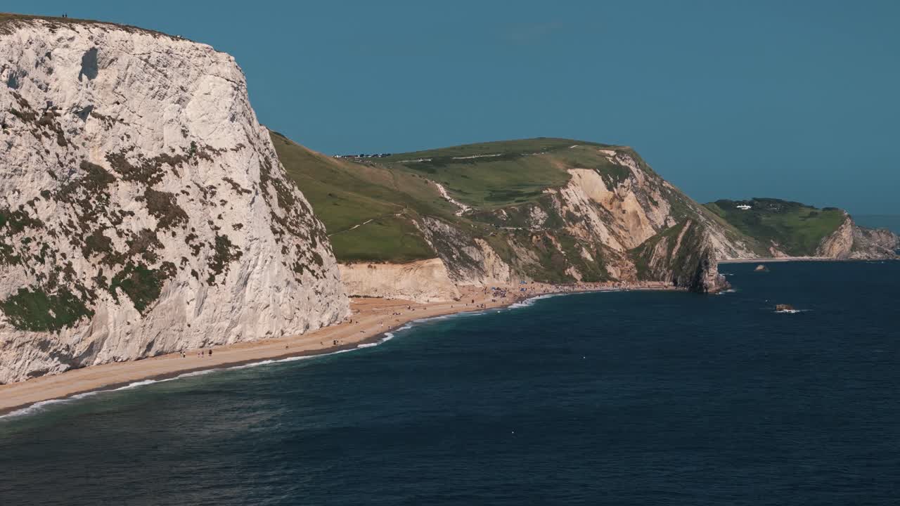 Slow approach drone flight toward Durdle Door with sea waves and beach unfolding ahead, establishing aerial