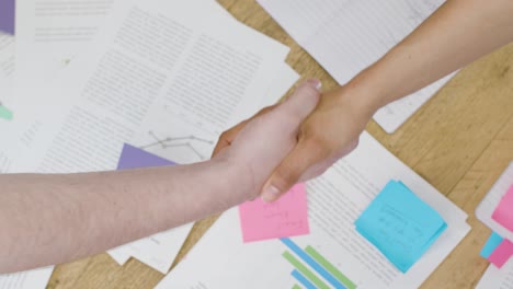 Overhead-Close-Up-Colleagues-Shaking-Hands-Over-Paperwork-On-Table