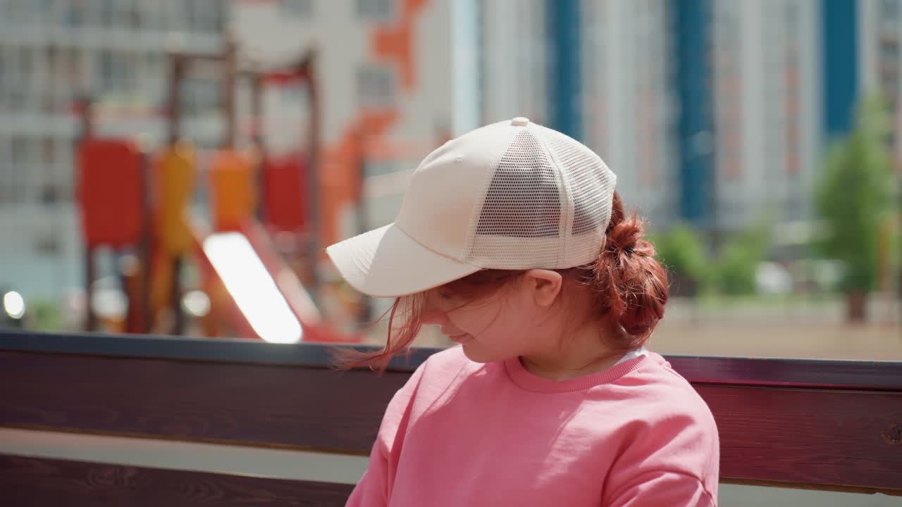 Closeup Portrait Of Teenager On Bench Wearing Cap And Pink Sweatshirt, Soft City Playground Bokeh Behind, Wind Tousled Hair And Subtle Smile, Calm Reflective Mood, Sunlight Highlights And Relaxed