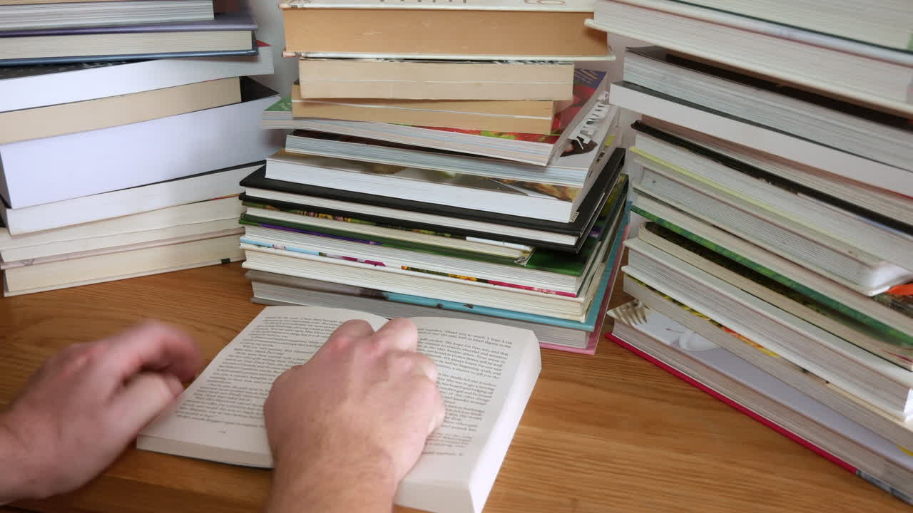 An unrecognizable man is reading a book with stacks of books on the desk