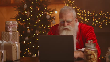 Papá-Noel-Real-Que-Utiliza-Nueva-Tecnología-Para-Comunicarse-Con-Los-Niños-Que-Reciben-Correo-O-Lista-De-Deseos.-Alegre-Trabajando-En-Una-Computadora-Portátil-Y-Sonriendo-Mientras-Está-Sentado-En-Su-Silla-Con-Chimenea-Y-árbol-De-Navidad.-Imágenes-4k-De-Alta-Calidad