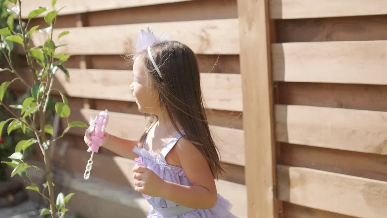 una niña con un vestido está soplando burbujas de jabón en el patio trasero en un soleado día de verano