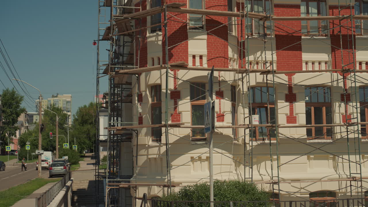 Facciata storica in mattoni illuminata dal sole e avvolta da impalcature, modanature decorate, balcone e cupola visibili, cavi elettrici aerei che attraversano un cielo azzurro limpido, lavori di conservazione e riparazione della muratura in corso, strada urbana