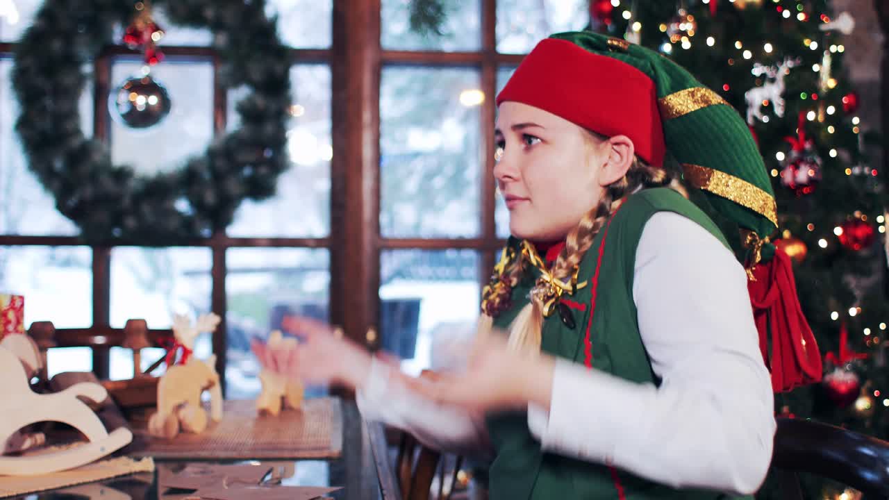 Elf girl sitting at the table with New Year's gifts chooses letters to Santa Claus. Room with large windows and christmas tree