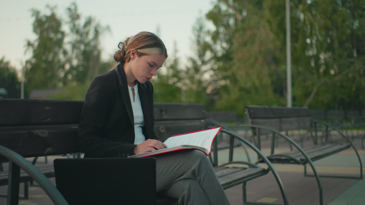 Hvit dame som sitter alene utendørs på en trebenk med en bærbar PC i nærheten i rolige urbane omgivelser omgitt av tomme benker, trær og en stolpe i bakgrunnen og leser nøye i boken sin