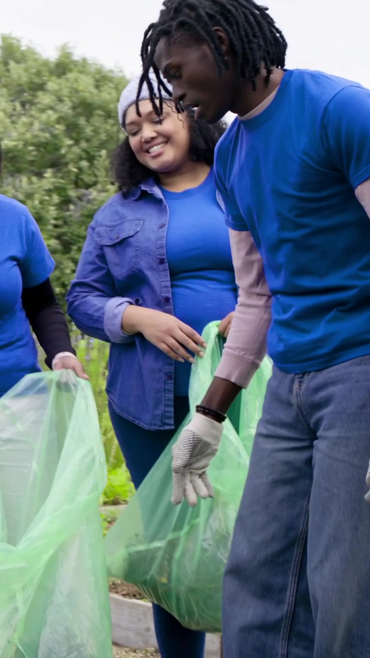 Vertical video: Holding green bag, woman in blue as gloved man picking debris for garden cleanup