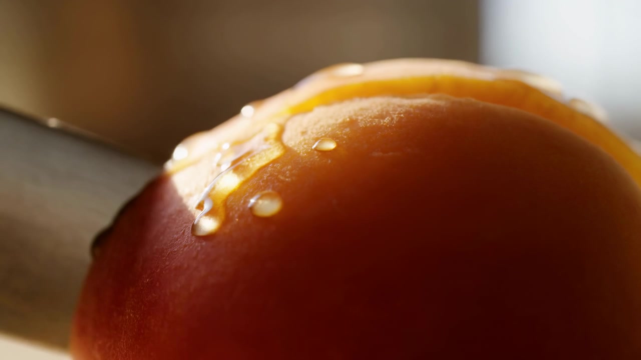 Primo piano di un pomodoro rosso maturo in basso a destra in primo piano mentre un coltello affetta da sinistra, con pelle lucida, goccioline e una cucina leggermente sfocata.