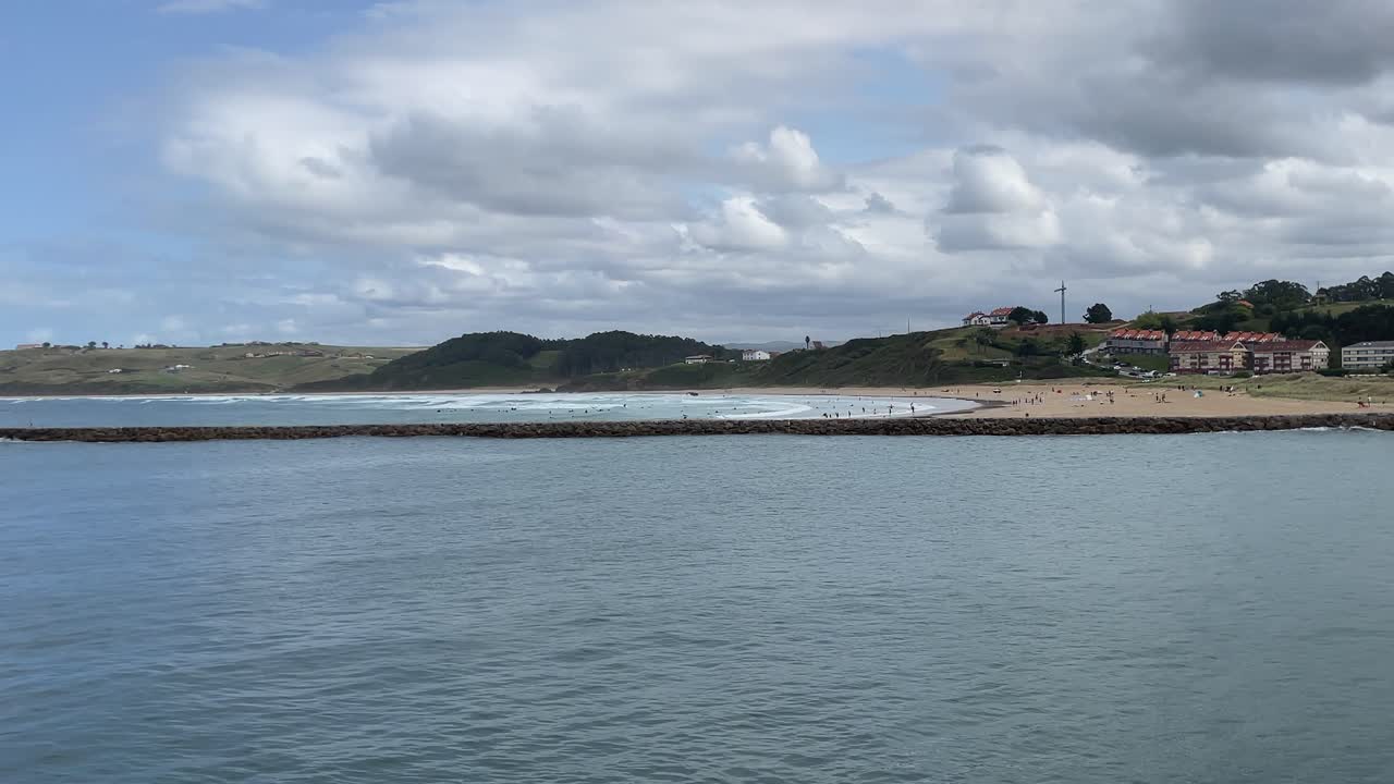 Filmagem em câmera lenta do mar ao lado de um quebra-mar de pedra, com a Playa de Merón no lado oposto em San Vicente de la Barquera. Um grande grupo de surfistas espera na água com suas pranchas.