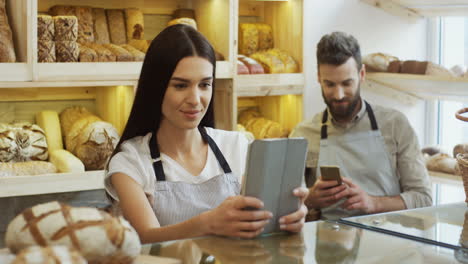 Pretty-Woman-Seller-Scrolling-And-Taping-On-The-Tablet-Computer-While-Standing-At-The-Counter-In-The-Bakery-Shop,-Man-Talking-On-The-Phone-Behind-Her