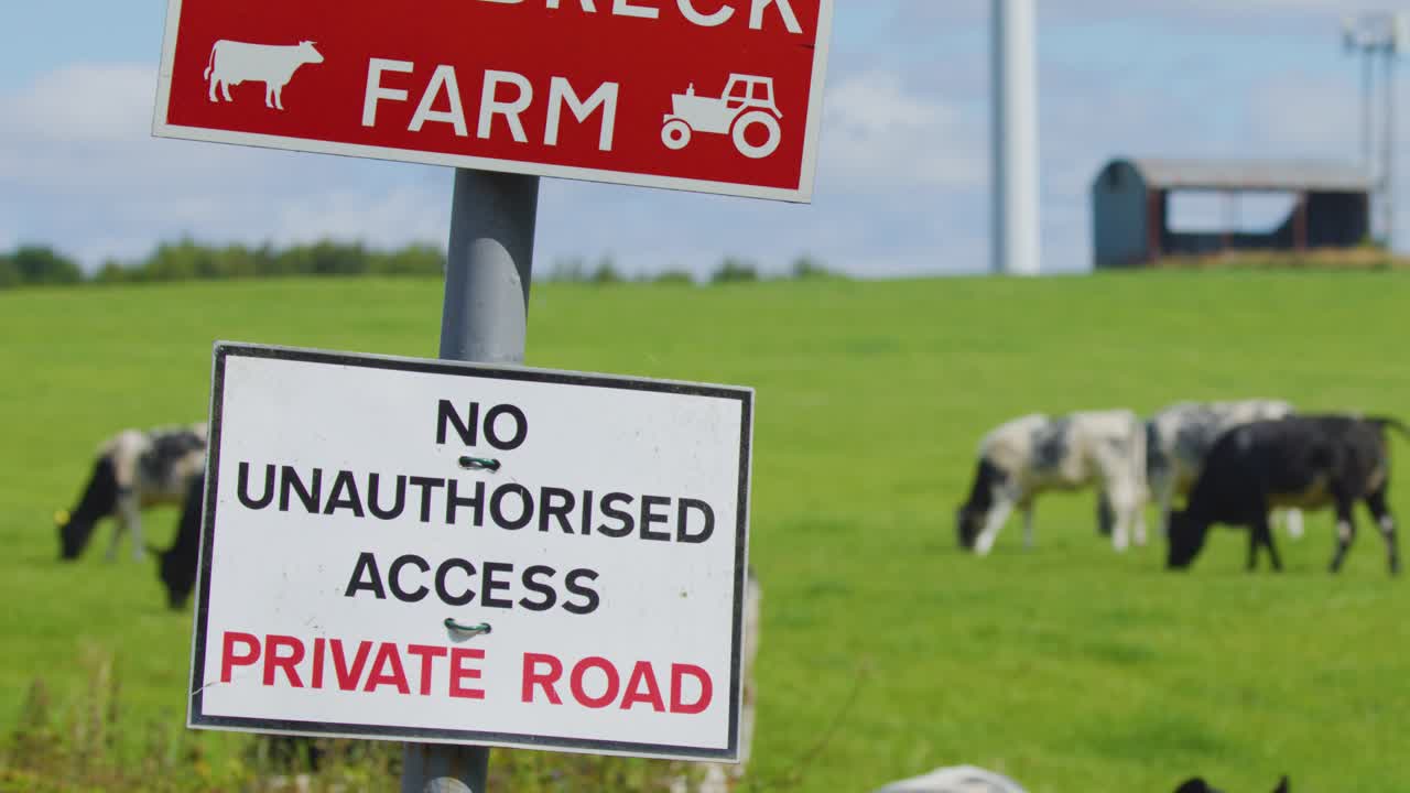 A warning sign marks a private farm road, with black and white cows grazing in a green field under daylight. Slight camera movement reveals rural landscape
