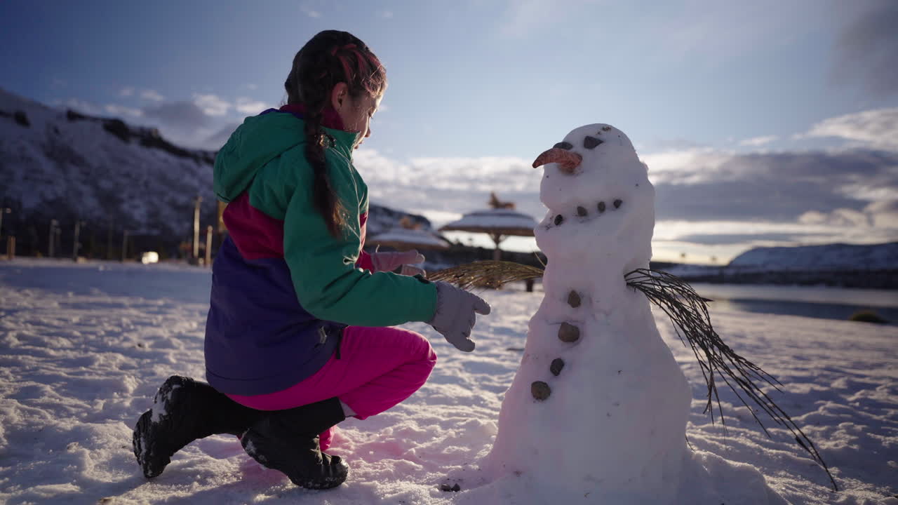 Child in colorful winter outfit adds stones to a snowman with carrot nose and twig arms, set against a bright sky and snowy mountains in Patagonia