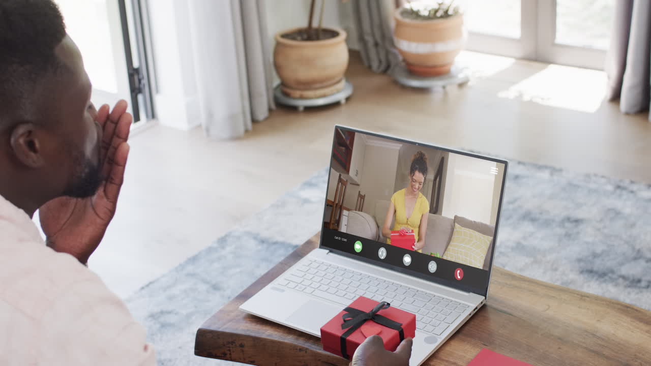 African american man holding red gift and using laptop with caucasian woman with gift on screen