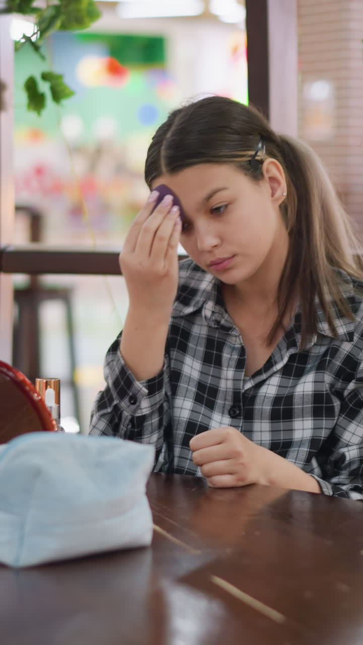 Adolescente probando una esponja de maquillaje en una zona de descanso concurrida de un centro comercial, fondo colorido y desenfocado, comprobando el tono bajo luces cálidas con un pequeño neceser y botes, momento espontáneo de aseo personal en público con actitud indecisa