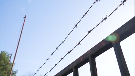 low-angle-view-POV-of-wire-fence-above-metal-doors-protective-layer-spikes-two-lines-from-entering-private-property-valuable-goods-inside-bright-day-sun-shine-optical-flare-high-pole-without-flag-tree