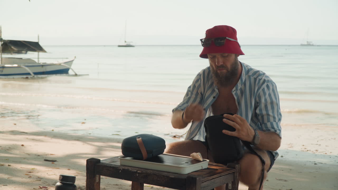 Cerimonia del tè in spiaggia con uomo caucasico che versa da una teiera su un tavolo di legno, che indossa un cappello da pescatore rosso, barche all'orizzonte, onde calme e tramonto caldo, viaggiatore solitario che prepara un rituale consapevole