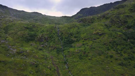 Vista-Aérea-Ascendente-De-Una-Cascada-Que-Fluye-Sobre-El-Borde-De-Una-Montaña-Cubierta-De-Hierba-En-Noruega