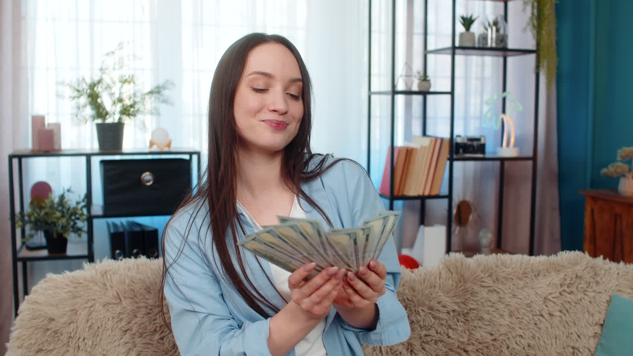 Mujer joven bailando alegremente en el sofá, sosteniendo billetes de dólar en efectivo y celebrando el éxito de haber ganado la lotería