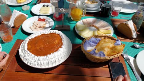eevated view family around a wooden table laden with various dishes, including a large cake-like dessert, pastries, scrambled eggs, and fruit and empanadas
