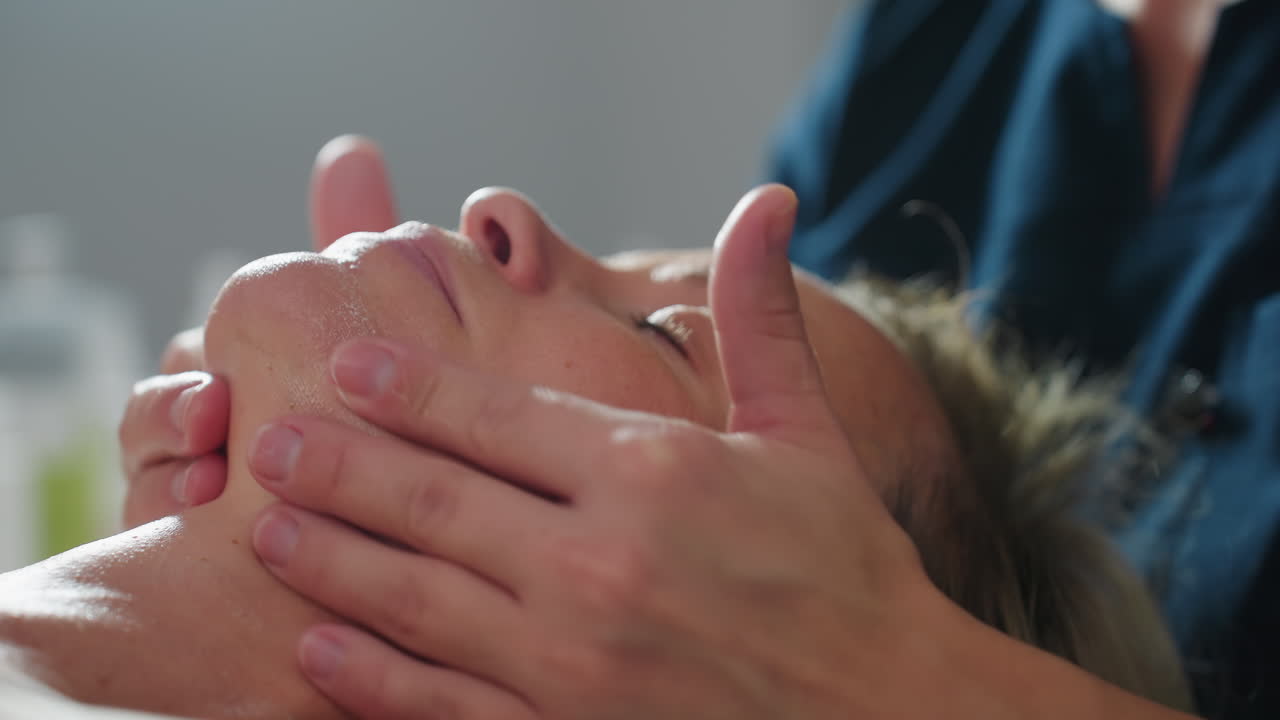 Female visitor lies relaxed with eyes closed while body work therapist gently massages product across cheeks and forehead, enhancing hydrated glow in tranquil spa treatment environment