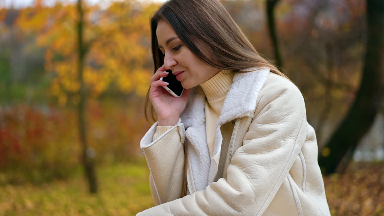 Brunette woman in warm clothes sitting on the table and speaking on the phone. Lady having phone conversation in the colorful autumn park.