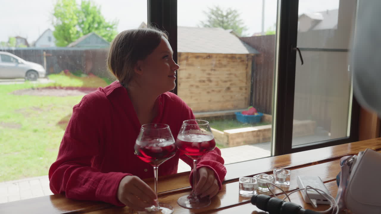Close up of woman sitting at table holding two glasses of red iced drinks while smiling at someone taking photo with phone, indoor setting with large window and backyard view in soft daylight