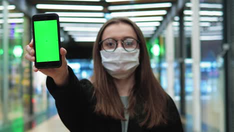 young woman stands in a medical mask and glasses. hold the phone with green screen. female smiling looking at camera. medical concept from coronavirus. close up portrait in 4k uhd 2160p