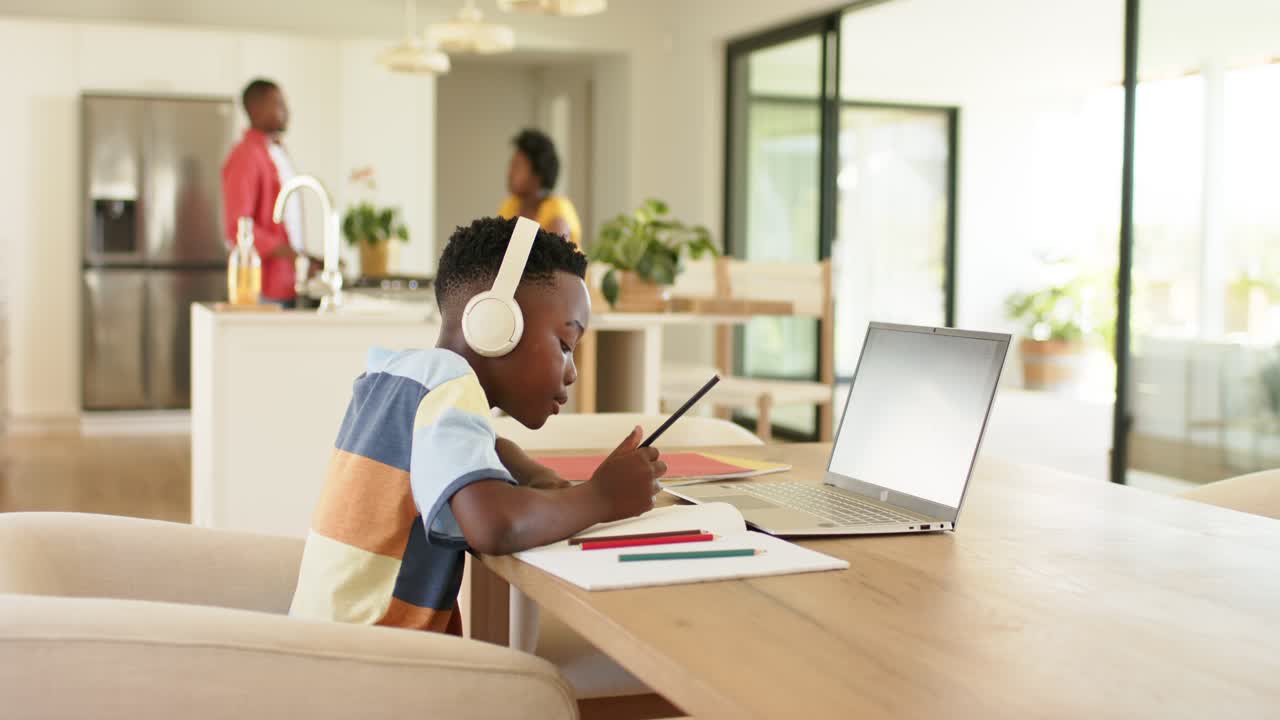African American boy listening on laptop with headphones at dining table, taking notes, copy space