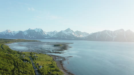 vista aérea sobre las casas y una carretera, hacia el mar poco profundo en el océano ártico y el pico nevado en el fondo, soleado, día de verano, en rotsund, troms, nordland, noruega - dolly, drone shot