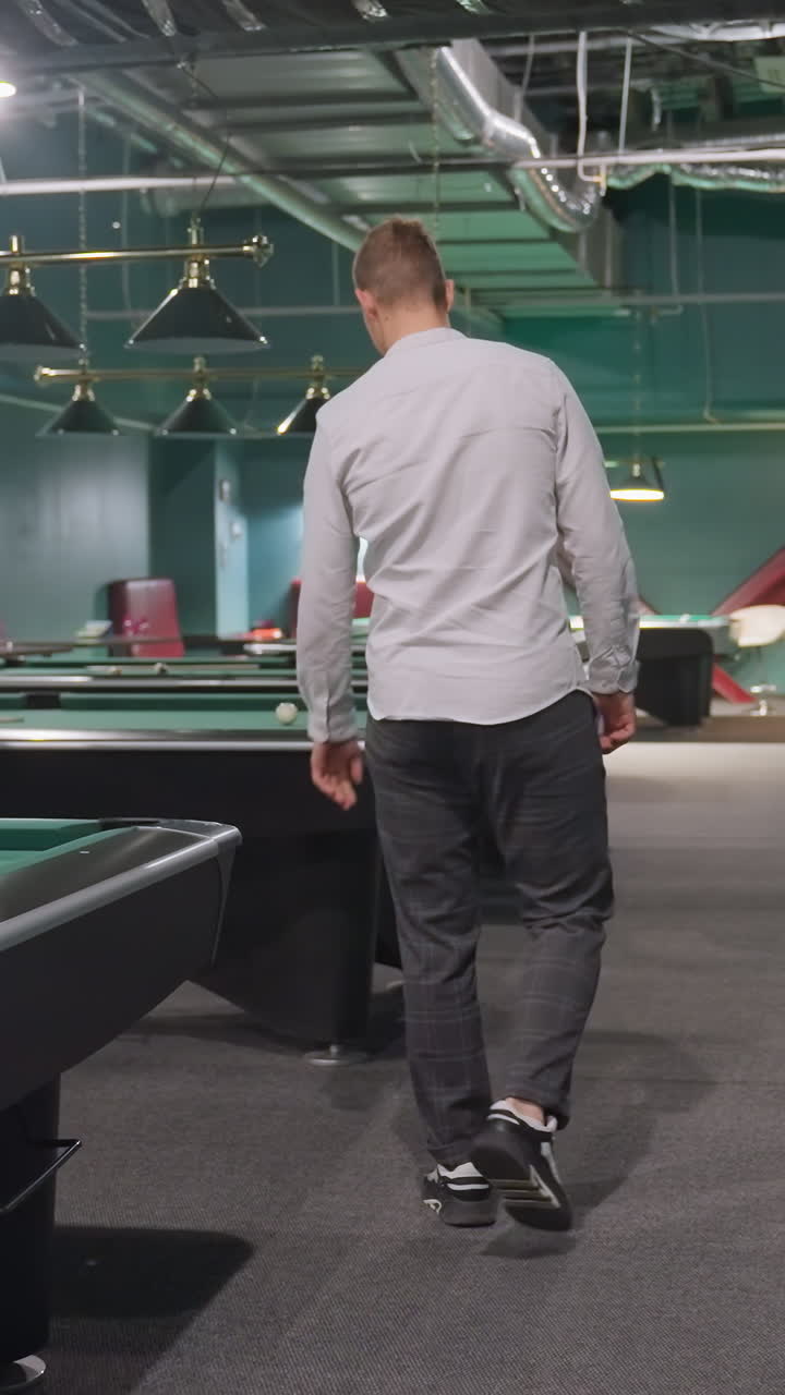 Foto da dietro di un giocatore con maglietta bianca e pantaloni grigi che entra in una sala da biliardo, fermo davanti a un tavolo da biliardo verde, che raccoglie una palla bianca. Ambiente moderno di un club sportivo con atmosfera di illuminazione dall'alto