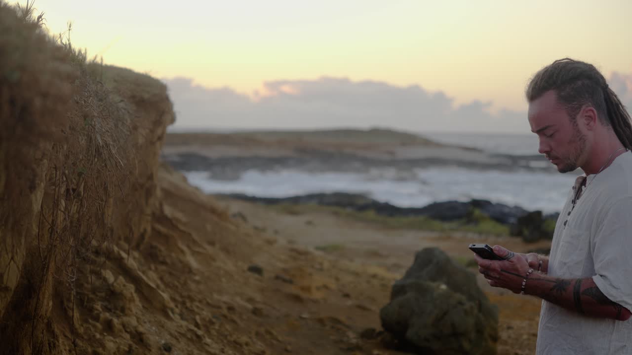 A man in white clothing stands in profile on a rocky coastal trail, looking down at his smartphone while ocean waves roll against the horizon under sunrise light