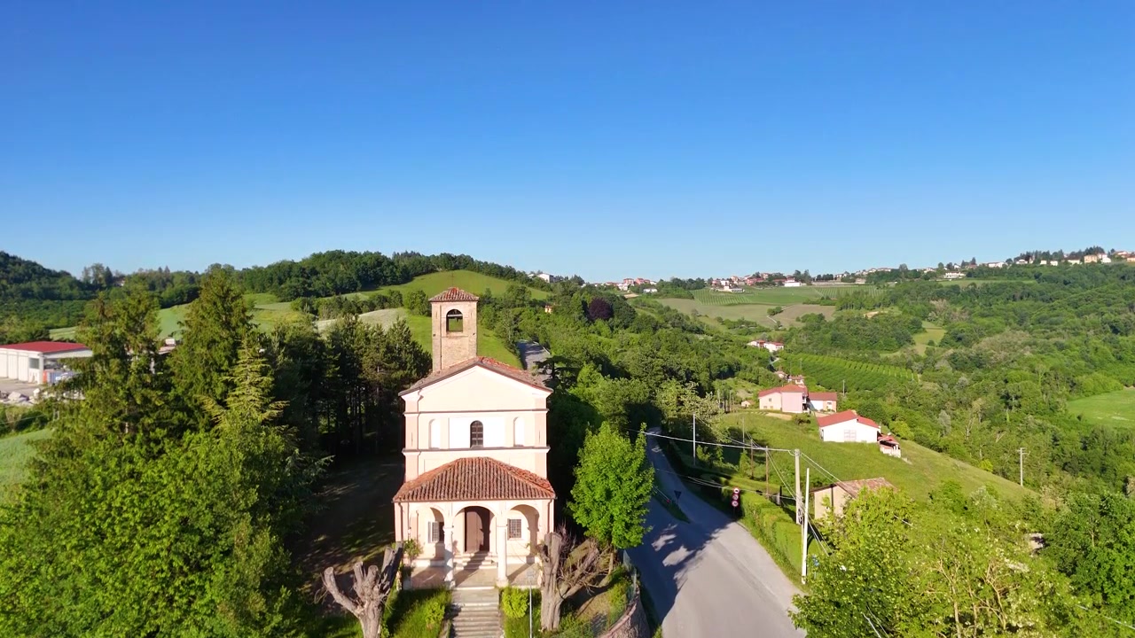 Una telecamera drone si alza sopra un prato di fiori selvatici verso una chiesa storica in Piemonte.