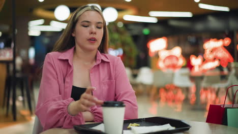 woman in pink dress enjoying coffee, placing the cup on a tray with leftover fries and tissue, rubbing her stomach with a satisfied expression with colorful blurred background and unclear items