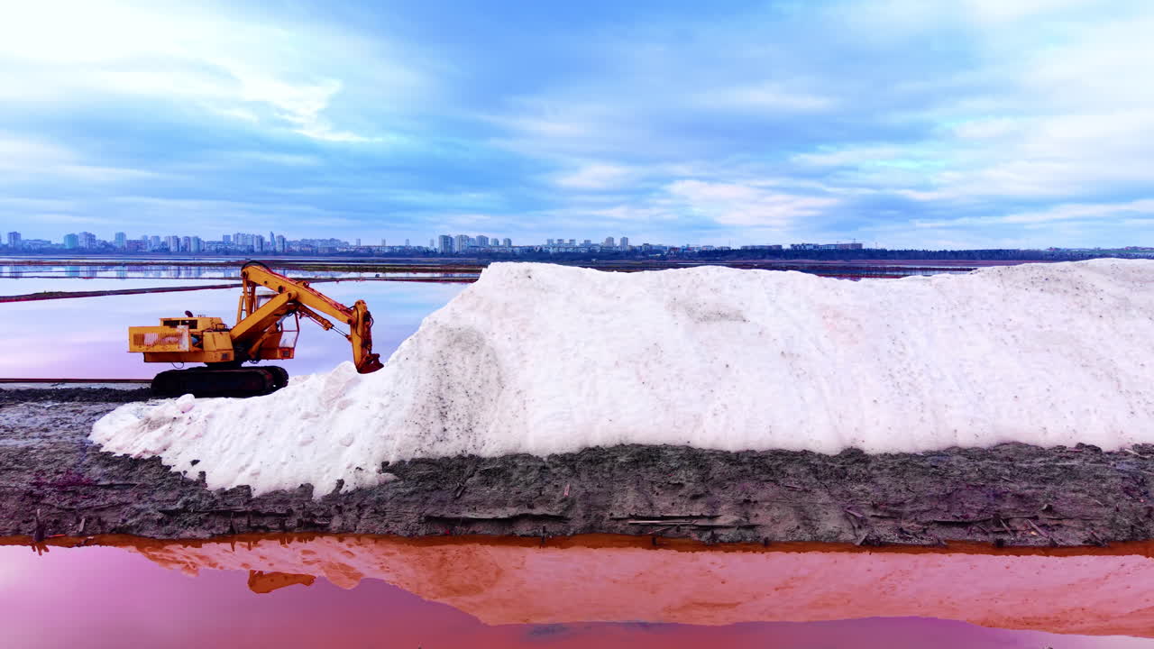 Vista laterale di un escavatore idraulico giallo su un argine di sale con edifici cittadini sullo sfondo e riflessi rosa sull'acqua. Escavatore industriale che lavora il sale marino in un sito di evaporazione.