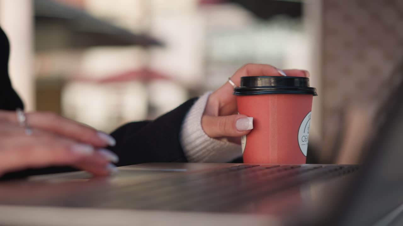 마우스를 사용하는 동안 노트북 근처에 커피 컵을 부드럽게 돌리는 여자의 손의 클로즈업, 작업 공간과 현대 환경의 흐릿한 배경과 함께 그녀의 매니큐어 손가락을 강조합니다.