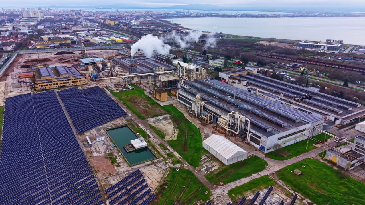 Solar panels and industrial buildings seen from above in a location near water with some clouds in the sky during the day