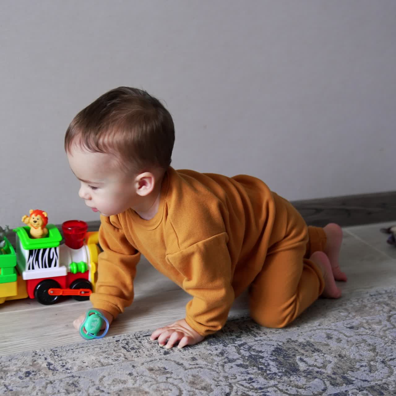 Beautiful baby boy in orange sport suit crawls by the floor holding a pacifier in his hand. Lovely kid trying to reach the desirable toy