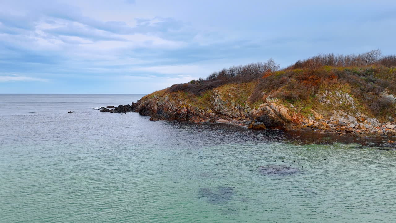 Vista della costa con acqua e rocce. Una costa rocciosa incontra acqua limpida sotto un cielo nuvoloso. La vegetazione costeggia la riva in un tranquillo ambiente esterno.