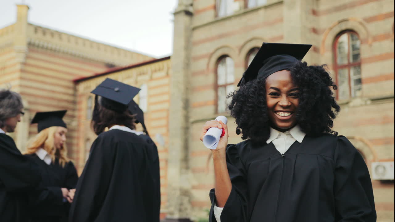Portrait of the African American happy young graduated woman posing to the camera, showing her diploma and making YES gesture in front of the University