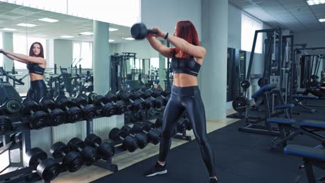 sportswoman in black top and leggings swinging hard kettlebell in fitness club. female athlete doing workout and looking at the mirror near rows of dumbbells.