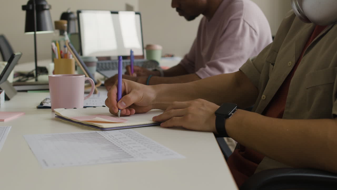 Two people working at a desk, writing and using a laptop